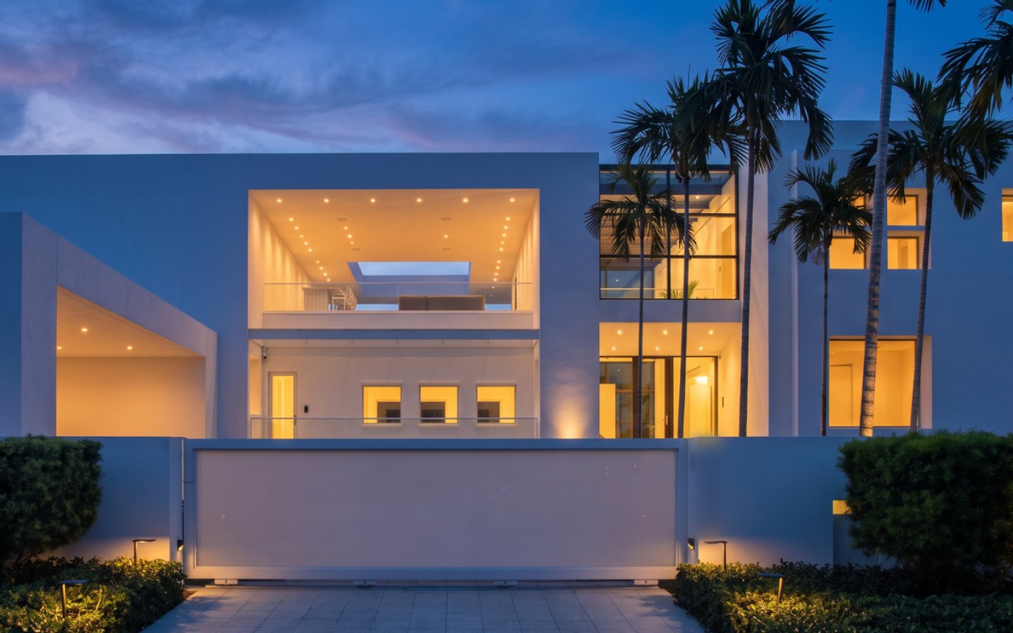 Modern white gated home after a day-to-twilight edit, with interior and landscape lights on